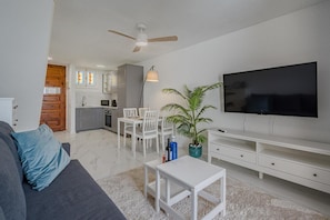 A modern living room with a sofa, TV, dining table, chairs, and a kitchen in the background.
