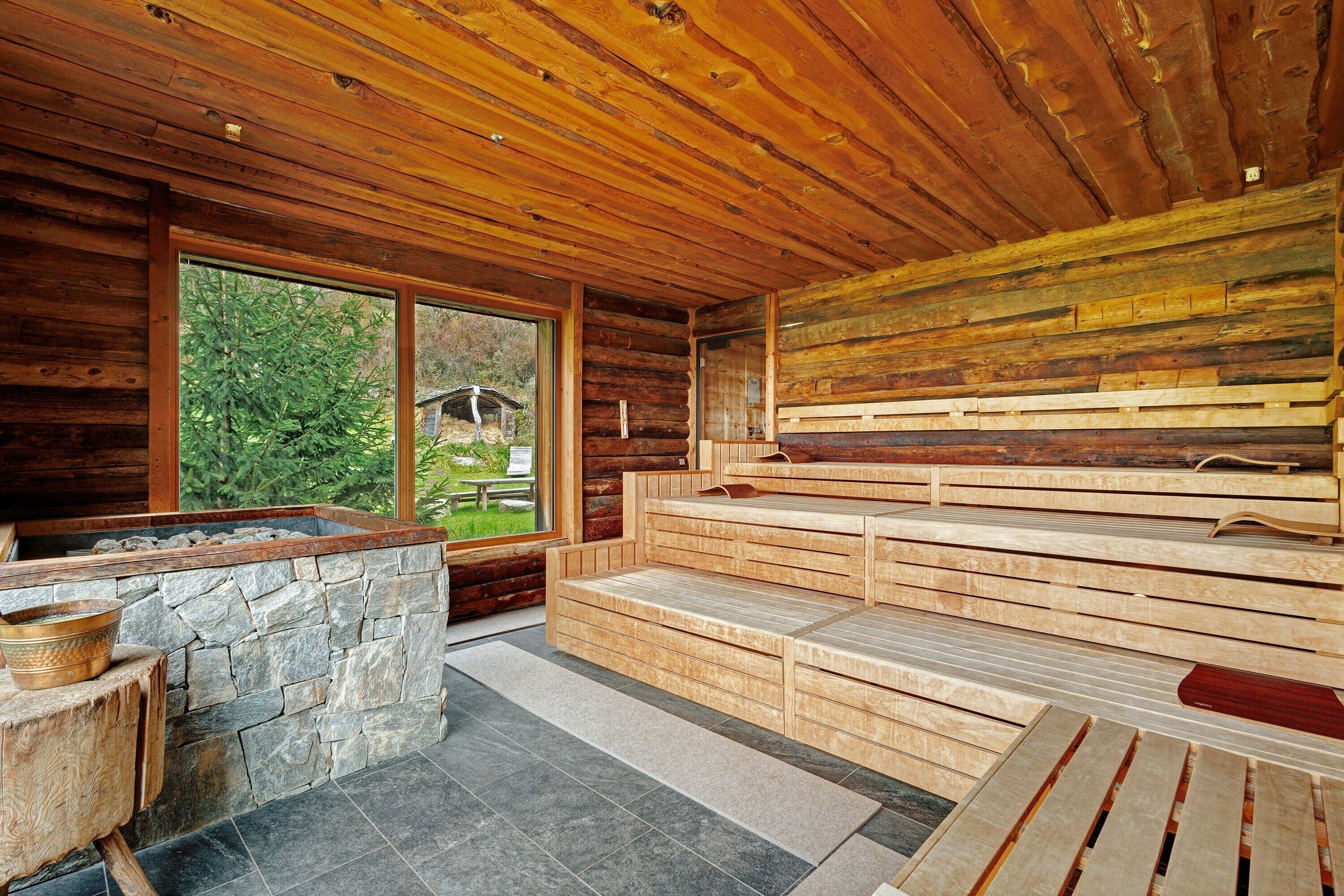 Innenansicht einer Holzhütte mit Kamin aus Stein, Holzbanken und einem großen Fenster mit Blick ins Freie.