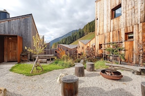 A rustic wooden building with a gravel courtyard, a small tree, and a fire pit.