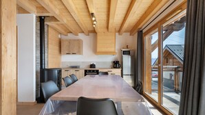 A wooden interior with a long dining table, chairs, a kitchen area, and large windows.