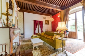A room with a green sofa, a white table, a patterned rug, a chandelier, and a wooden ceiling.