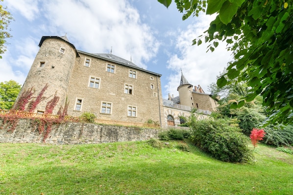 A historic stone building with multiple windows and a tower, surrounded by greenery and a clear sky.