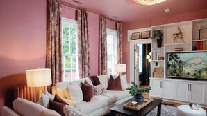 A living room with a white sofa, a coffee table, and a television. There are curtains on the windows, and a bookshelf with various items. - Downham House: Spacious 4BR, Old Town ALX (Alexandria)