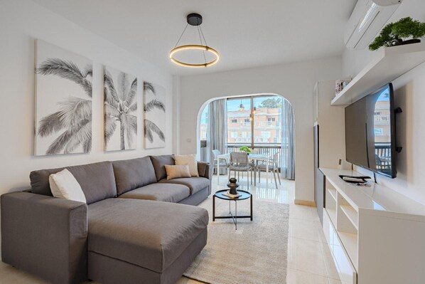 A modern living room with a grey sofa, a coffee table, and a flat-screen TV on a white console. There are wall art pieces and a large window with a view of buildings.