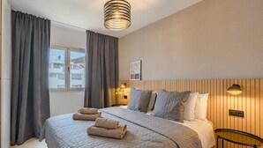 A bedroom with a bed, curtains, a bedside table, a wall-mounted lamp, and a window.