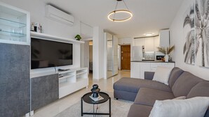 A modern living room with a flat-screen TV, a grey sofa, a coffee table, and a kitchen in the background.