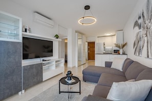 A modern living room with a flat-screen TV, a grey sofa, a coffee table, and a kitchen in the background.