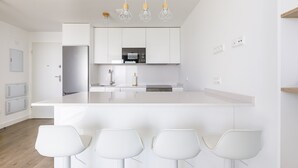 Eine moderne Küche mit weißen Schränken, einem Edelstahl-Kühlschrank, einer Mikrowelle und einem eingebauten Backofen. Vor der Arbeitsplatte stehen vier weiße Barhocker.