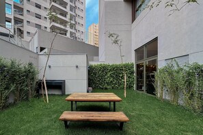 A modern outdoor patio with a wooden table and bench, surrounded by a well-maintained hedge and a grassy area. - Temporada Express (São Paulo)