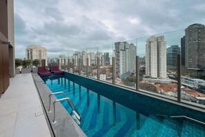 Ein Pool auf dem Dach mit Blick auf die Stadt, Glasgeländern und roten Liegestühlen.