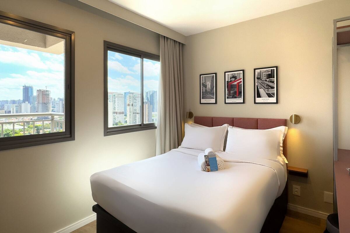 Ein Hotelzimmer mit einem großen Bett, Blick auf die Stadt und drei gerahmten Bildern an der Wand.