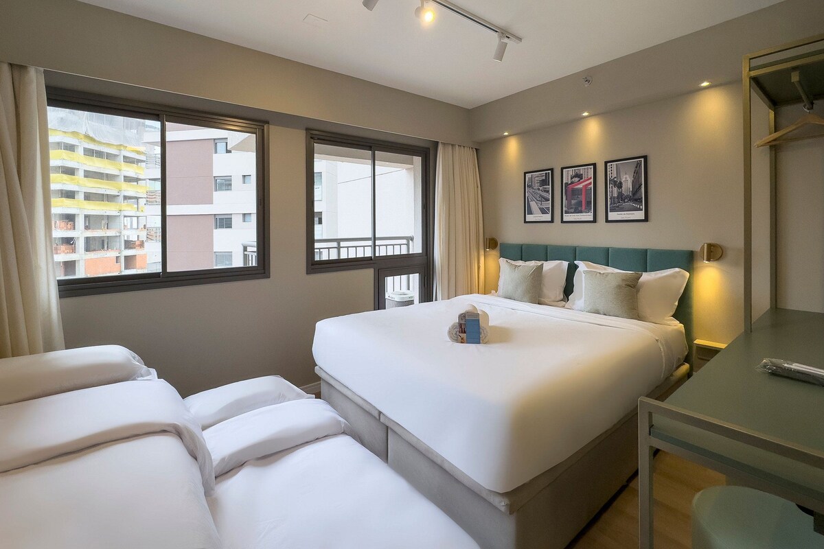 A hotel room with a large bed, two bedside tables, a desk, and a window with a view of buildings.