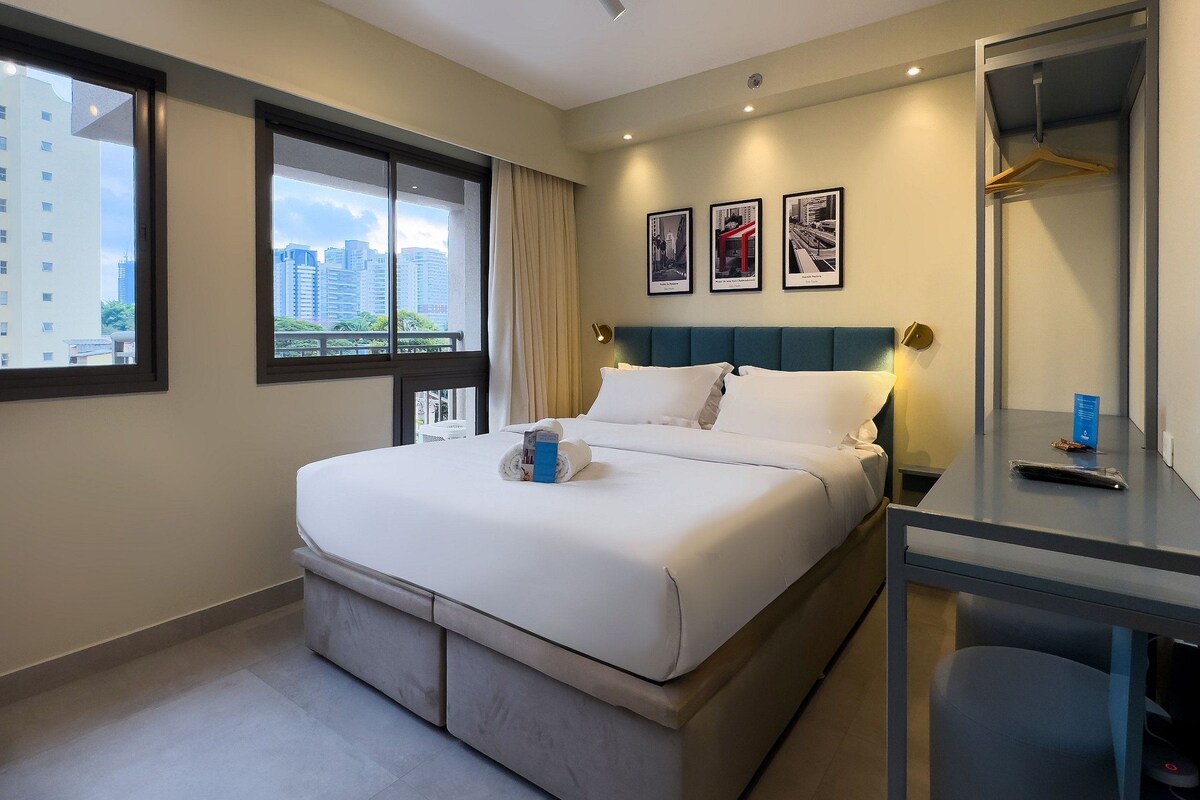 A hotel room with a bed, a desk, a chair, a window with a view of buildings, and a wall with framed pictures.