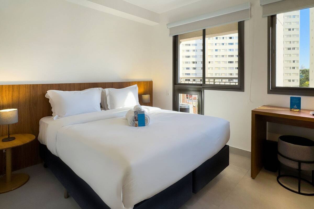Ein Hotelzimmer mit einem großen Bett, einem Holzkopfende, einem Schreibtisch mit Stuhl und einem Fenster mit Blick auf hohe Gebäude.
