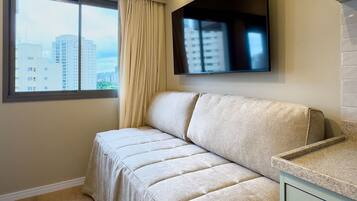 A modern hotel room with a sofa, a flat-screen TV mounted on the wall, a window with a view of buildings, and a ceiling fan.