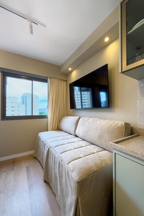 Ein modernes Hotelzimmer mit einer Couch, einem an der Wand befestigten Flachbildfernseher, einem Fenster mit Blick auf Gebäude und einem Deckenventilator.