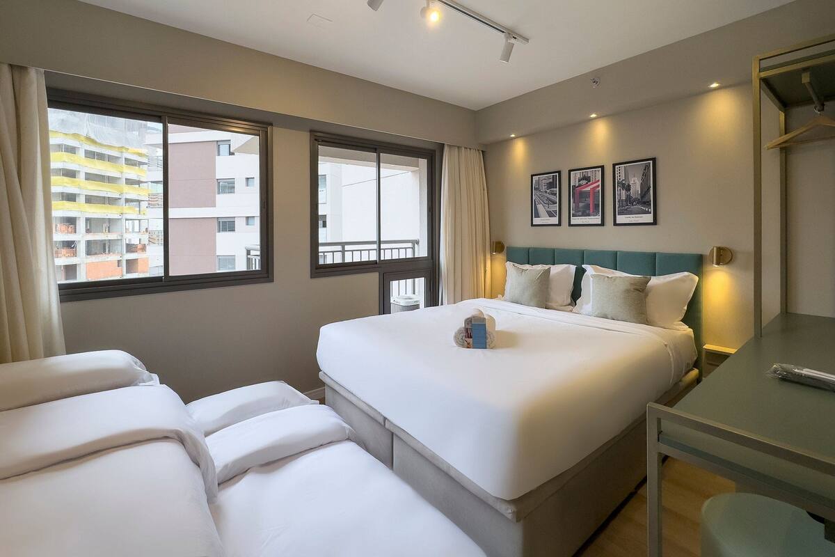 Ein Hotelzimmer mit einem Bett, zwei Nachttischen, einem Schreibtisch und einem Fenster mit Blick auf Gebäude.