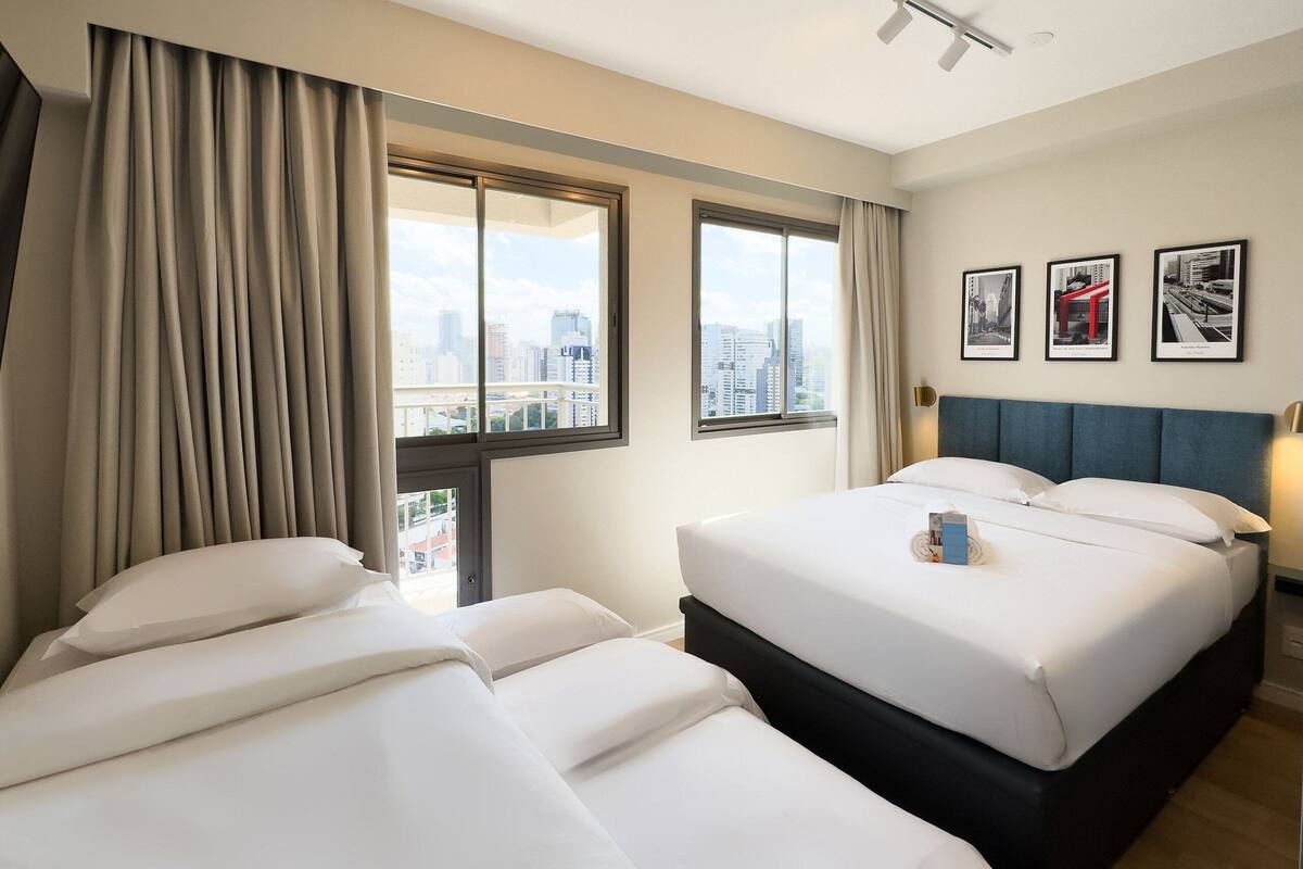Ein Hotelzimmer mit zwei Betten, einem großen Fenster mit Blick auf die Stadt und drei gerahmten Bildern an der Wand.