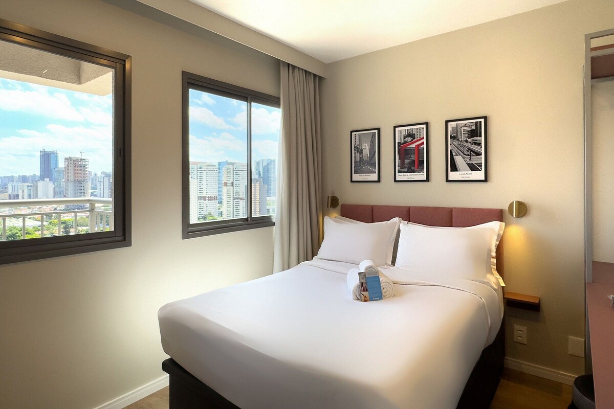 A hotel room with a large bed, a city view, and three framed pictures on the wall.