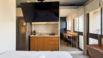 A modern hotel room with a large bed, a wooden kitchenette, a dining area, and a view through large windows.