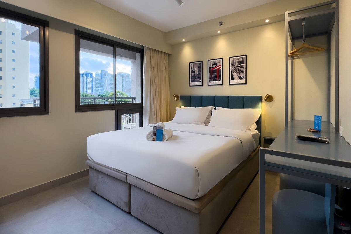 Ein Hotelzimmer mit Bett, Schreibtisch, Sessel, Fenster mit Blick auf Gebäude und Wand mit gerahmten Bildern.