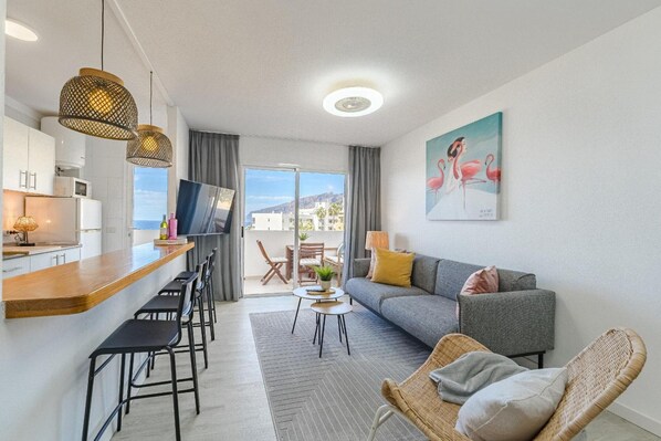 A modern hotel room with a kitchen area, a sofa, a dining table, and a view of the outdoors.
