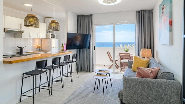 A modern kitchen with a flat-screen TV, a dining area with bar stools, a sofa, and a balcony with a view. - Vacation Apartment 'Playa La Arena 2 Bed' with Sea View, Balcony and Wi-Fi (Puerto de Santiago)