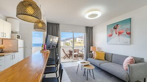 A modern room with a wooden dining table, a grey sofa, a TV, and a painting of flamingos. - Vacation Apartment 'Playa La Arena 2 Bed' with Sea View, Balcony and Wi-Fi (Puerto de Santiago)