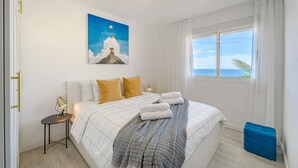 A bedroom with a bed, a nightstand, a painting, a window with curtains, and a view of the sea. - Vacation Apartment 'Playa La Arena 2 Bed' with Sea View, Balcony and Wi-Fi (Puerto de Santiago)