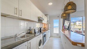 A modern kitchen with white cabinets, a built-in washing machine, and a view of the city through large windows. - Vacation Apartment 'Playa La Arena 2 Bed' with Sea View, Balcony and Wi-Fi (Puerto de Santiago)