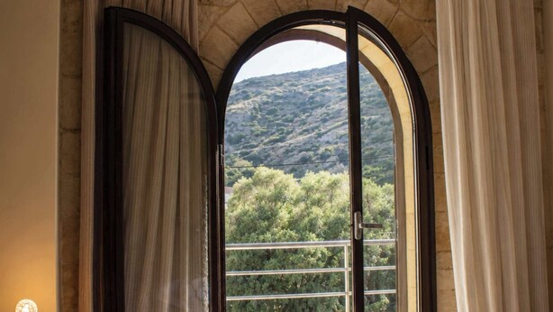 A bedroom with a bed, pillows, a bedside table, and a view of a mountainous landscape through an arched window. - Xristinas Apartments - Artemis (Kolymvari)