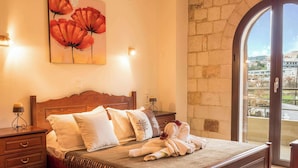 A bedroom with a wooden bed, multiple pillows, a nightstand, a lamp, and a painting of red flowers on the wall. - Xristinas Apartments - Artemis (Kolymvari)