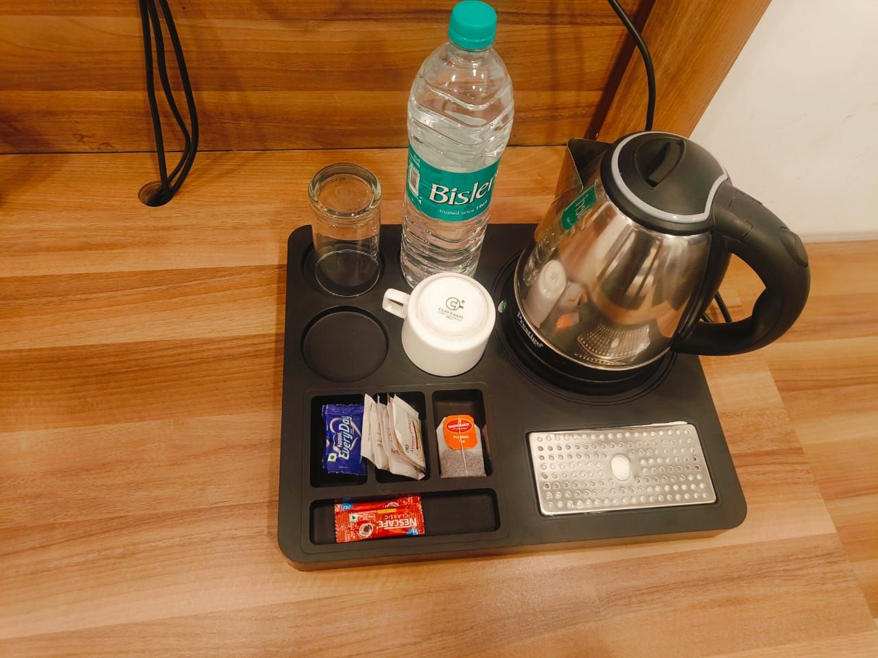 A tray with a Bisleri water bottle, a glass, a white mug, a stainless steel kettle, and packets of snacks.