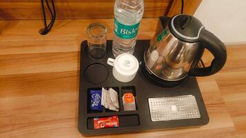 A tray with a Bisleri water bottle, a glass, a white mug, a stainless steel kettle, and packets of snacks.