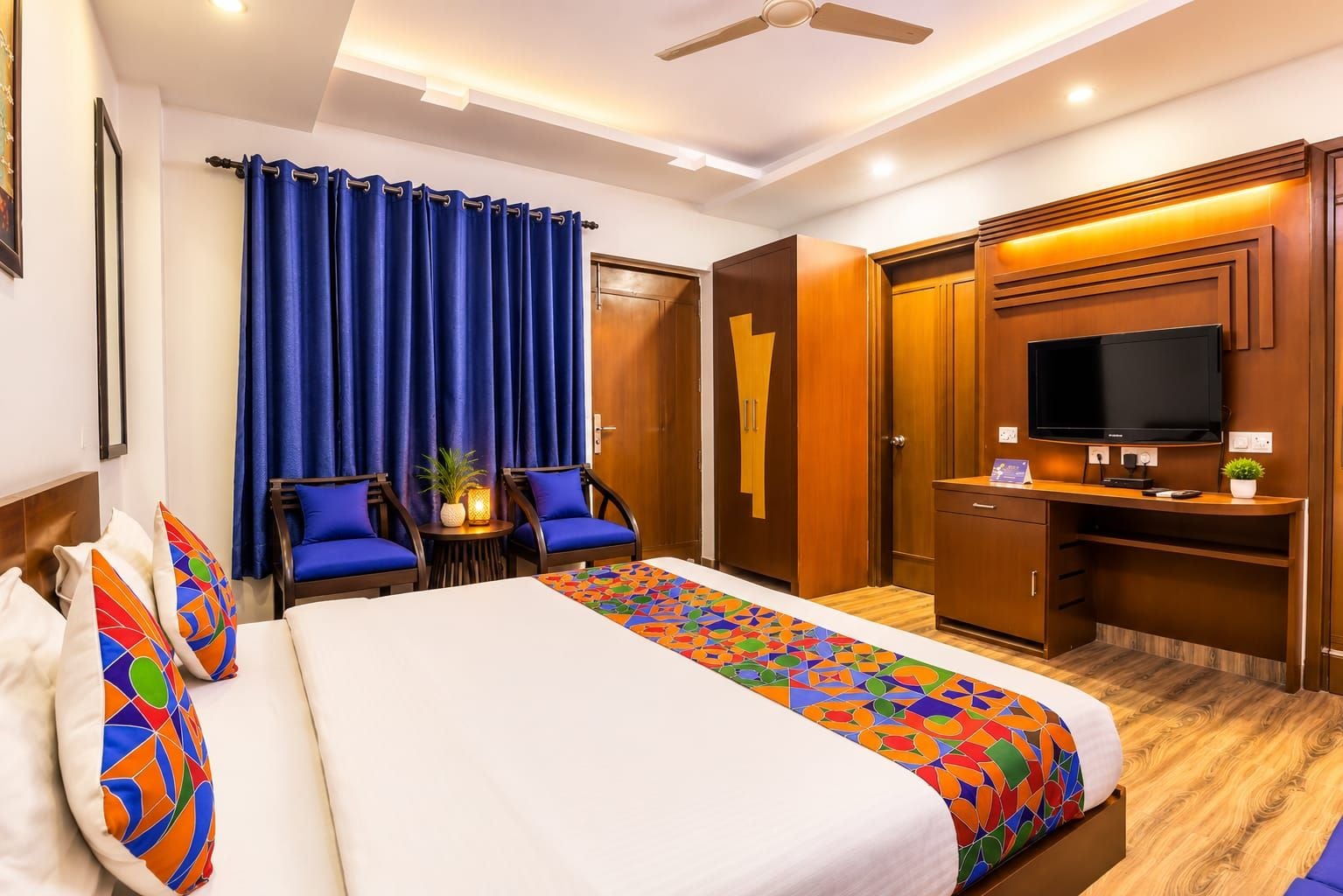A hotel room with a bed, two chairs, a desk with a TV, and a wardrobe.