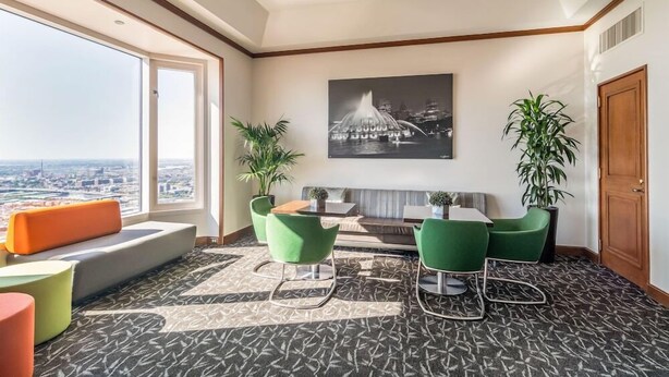 A modern hotel room with a large window, a sofa, green chairs, a small table, and a potted plant. - The Buckingham Athletic Club & Hotel (Chicago)