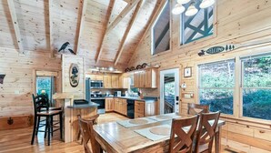 A wooden cabin interior with a dining area, kitchen, and large windows offering a view of the forest. - Luxury Cabin w/ Spa, Firepit & Game Room (Blue Ridge)
