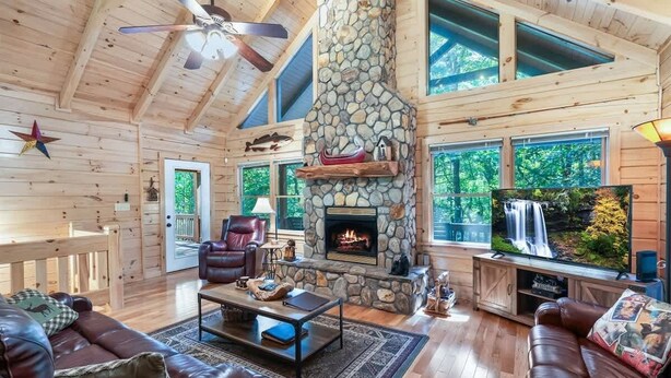 A log cabin interior with a stone fireplace, leather furniture, and a large window overlooking a waterfall. - Luxury Cabin w/ Spa, Firepit & Game Room (Blue Ridge)