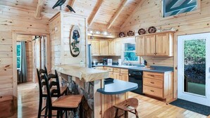 A wooden cabin interior with a kitchen, dining area, and a stone fireplace. - Luxury Cabin w/ Spa, Firepit & Game Room (Blue Ridge)