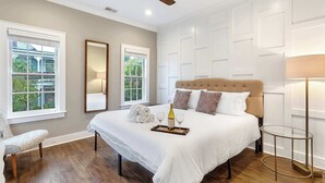 A bedroom with a large bed, a ceiling fan, two windows, a bedside table, and a lamp.