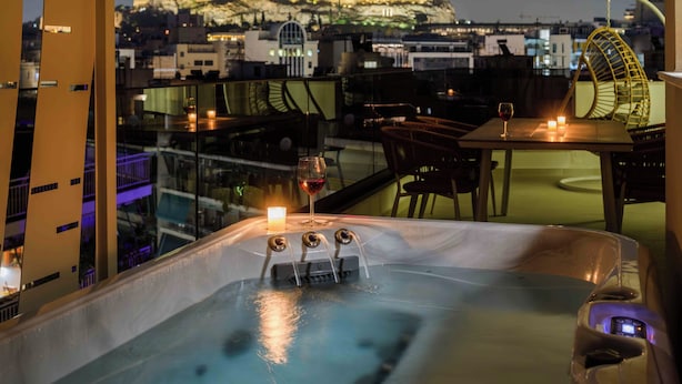 A hot tub with a glass partition, a candle, and a glass of red wine on a rooftop terrace with a city view. - Thea Acropolis Suites & Apartments (Athens)