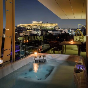 Un jacuzzi con separador de vidrio, una vela y una copa de vino tinto en una terraza en la azotea con vista a la ciudad.