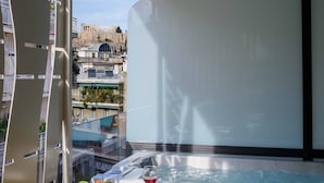 A balcony with a hot tub, a table with fruits and a glass of red wine, and a view of historic buildings. - Thea Acropolis Suites & Apartments (Athens)