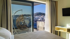 A hotel room with a bed, a hanging chair, a TV, and a view of a cityscape through a large window. - Thea Acropolis Suites & Apartments (Athens)