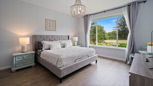 Een slaapkamer met een groot bed, twee nachtkastjes, een kroonluchter, een raam met gordijnen en uitzicht op groen.