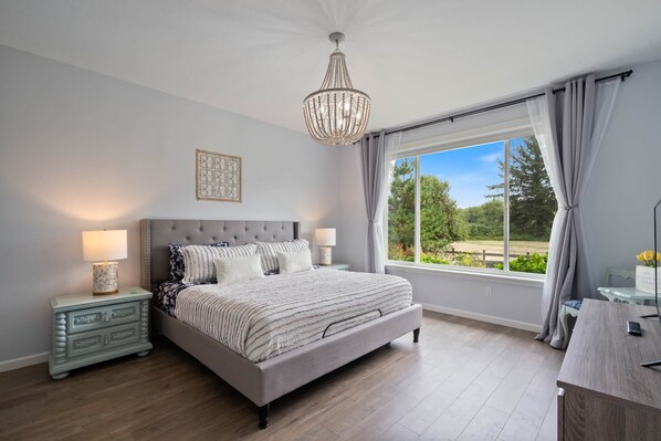 Une chambre avec un grand lit, deux tables de chevet, un lustre, une fenêtre avec des rideaux et une vue sur la verdure.