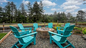 Een ronde vuurkuil omringd door vier turquoise Adirondack-stoelen in een grindgebied, met een houten omheining en bomen op de achtergrond.