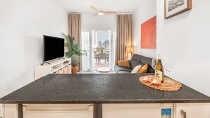 Una cocina moderna con una televisión de pantalla plana, un comedor con mesa y sillas, y una sala de estar con sofá e imagen enmarcada en la pared.