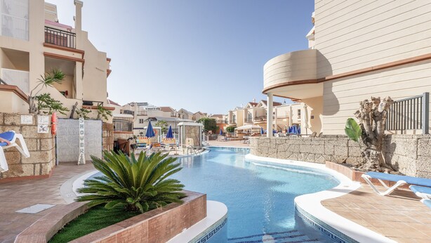 Una piscina de borde curvo, rodeada de sillones de descanso y sombrillas, con edificios al fondo.