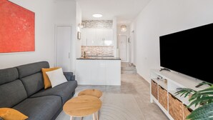 Una sala de estar moderna con un sofá gris, mesitas de madera, un televisor de pantalla plana y una cocina al fondo.